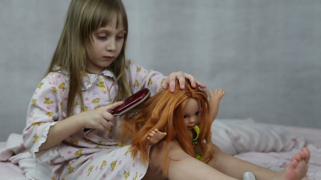 Girl playing with doll sitting on the bed, she braids her hair

