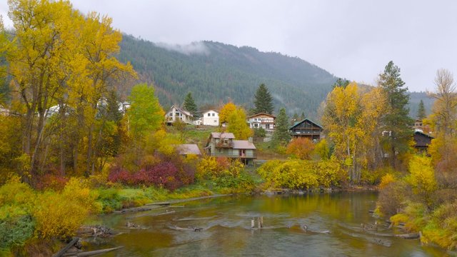 Autumn forest on the riverside, beautiful trees and small houses on a background of mountains