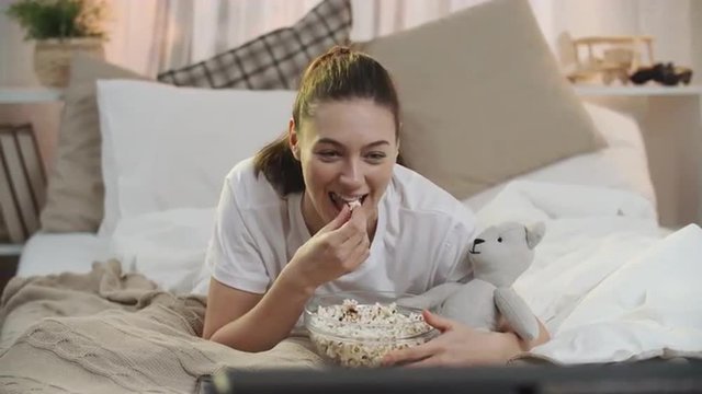 Pretty young woman lying in her bed with teddy bear, watching movie and laughing