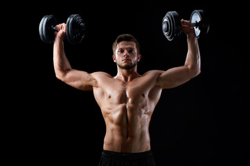 Naklejka na ściany i meble Reaching out for goals. Young serious muscular handsome man holding up two dumbbells exercising on black background