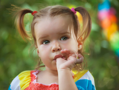 cute little girl wearing colorful dress sucks her thumb. Bad habit. in the park