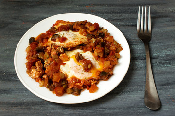 Shakshuka and fork on gray blue background. Two eggs in tomato sauce with vegetables.
