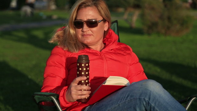 Middle age, blonde  woman reading a book  Sitting on lounge chair and dringing coffee at thermos. Resting and relaxing at the park