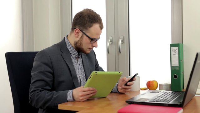 Young men in suit sitting in the office, behind desk ane rewriting something from tablet into phone. Positive emotions