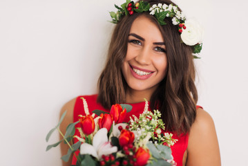 beautiful young girl in a red dress with a wreath on his head and a bouquet of flowers on a white background looking at the camera