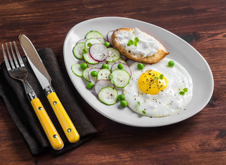 Fried egg, salad with cucumbers, radishes and green peas, toast with feta cheese on a light ceramic plate on dark wooden background. Healthy breakfast