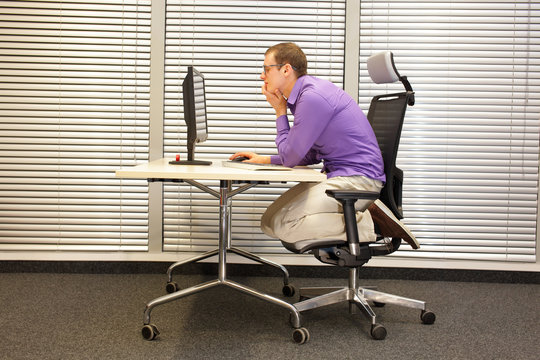 text neck - man in slouching position kneeling on ergonomic chair working with computer at desk