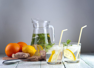 Glasses of detox drink with mineral water, lemon and mint, glass jug, lemon, orange and ginger on wooden table. Drink for diet. Focus on glasses.