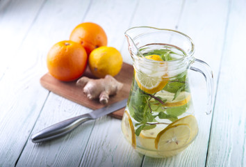 Glass jug with mineral water, lemon slices and mint leaves, ginger, lemon and oranges with knife on wooden table. Drink for diet. Focus on jug.