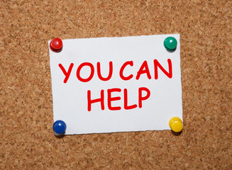 The phrase You Can Help  typed on a white piece of note paper and pinned to a cork notice board