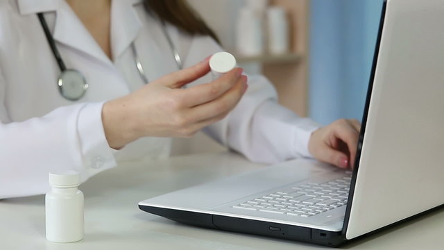 Female M.D. in white coat using laptop, typing online prescription for patient