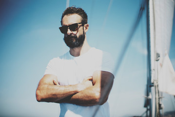 Fototapeta premium Portrait of young bearded man standing on the yacht, wearing sunglasses in sunny day. his arms are crossed. Horizontal mockup