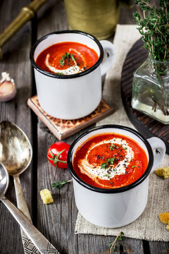Tomato cream soup in mugs and greens on a wooden table.