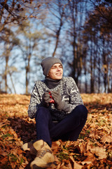 Handsome stylish happy young man in the sweater and hat with the cup of tea or coffee in the autumn park