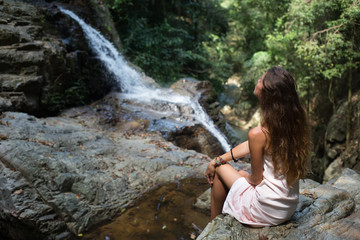 The beautiful fashionable girl with long hair, sits on surface stones, dressed in a light white dress, finding on tropical falls of the island Samui. Sunny day and happy smile. 