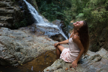 The beautiful fashionable girl with long hair, sits on surface stones, dressed in a light white dress, finding on tropical falls of the island Samui. Sunny day and happy smile.