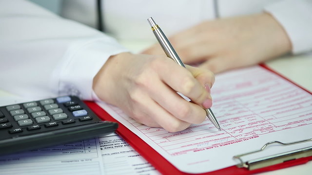 Closeup of doctor's hands filling in health insurance claim form, checking costs