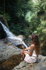 Fototapeta na wymiar The beautiful fashionable girl with long hair, sits on surface stones, dressed in a light white dress, finding on tropical falls of the island Samui. Sunny day and happy smile.