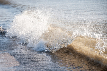 Eine sonnendurchflutete Welle bricht am Ufer. Tropfen fliegen und die Gischt sprüht. 