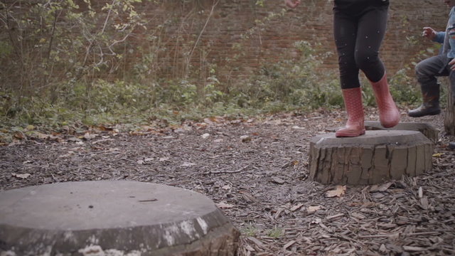 Children with wellies hopping on stepping stone logs in outdoor play area.