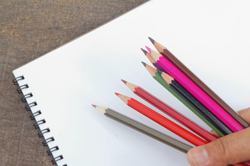 Blank notepad on the wooden table and a hand holding colorful color pencils. View from above