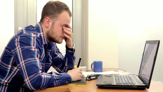 Young men in colrfull shirt, sitting behind his desk and doing something on his computer, he is tired and mad because of his work to do.