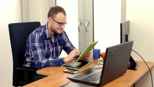 Young handsome men, siting behing his desk and alternately using tablet and smartphone, he is smiling. Positive emotions