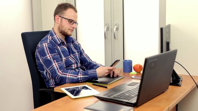 Young men in colorfull shirt sitting behing his computer, picks up the phone and speaking with someone. During conversation he is drinking from blue cup.