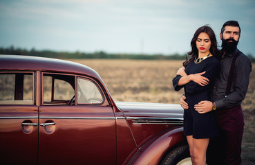 brutal bearded man with a mustache in a shirt, pants with suspenders with a girl with dark hair and big lips with bright red lipstick in a short dress and heels near retro car at sunset