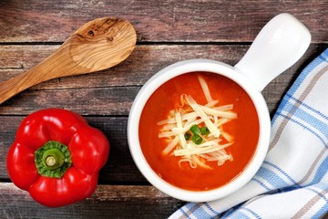 Red pepper soup topped with shredded cheese and green onions, overhead scene on rustic wood background