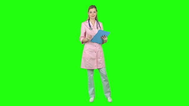 Beautiful young nurse standing on green screen background, writing something then looking at camera and smiling