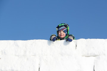 Fototapeta na wymiar Happy child in a color jacket with a hood at wall top from snow blocks 