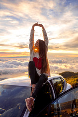 Young woman in sweater with heart shape rising hands sitting on the car roof above the clouds on...