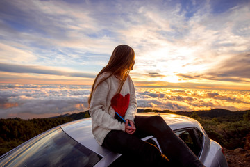 Fototapeta premium Young woman in sweater with heart shape enjoying beautiful cloudscape sitting on the car roof above the clouds on the sunrise