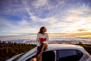 Naklejka premium Young woman in sweater with heart shape enjoying beautiful cloudscape sitting on the car roof above the clouds on the sunrise