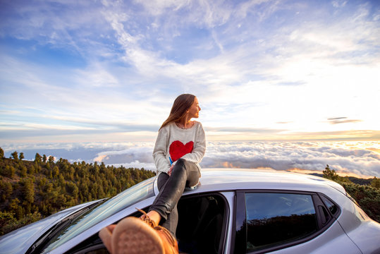 Young woman in sweater with heart shape enjoying beautiful cloudscape sitting on the car roof above the clouds on the sunrise