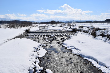 Fototapeta na wymiar 小川の雪溶け／冬は長い期間、鉛色の雲に被われる雪国の山形県ですが、春が近くなり、久し振りの好天で雪が溶け始めました。小川の雪溶け水がせせらぐ風景を撮影した、春の息吹を感じる早春イメージの写真です。