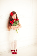 Obraz na płótnie Canvas Beautiful baby girl 4-5 year old standing in room holding red tulips. Looking at camera. Mothers day. Childhood. Romance.
