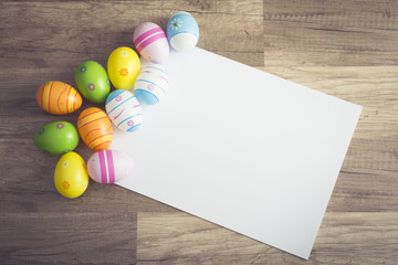 Photograph of a couple of colorful easter eggs  alligned around a sheet of white paper with copy-space on wooden surface