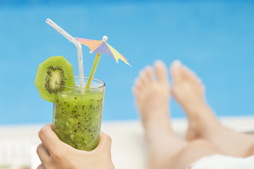Unrecognizable young female laying on sunbed during the sunny day and holding a glass of fresh kiwi smoothie with nice decoration at the poolside.