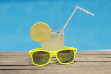 Glass of fresh lemonade with drinking straw and garish green sunglasses on the table. Swimming pool blurred background.