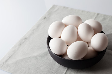 White eggs in a black bowl on beige off-white linen napkin on white background from side off centre composition  