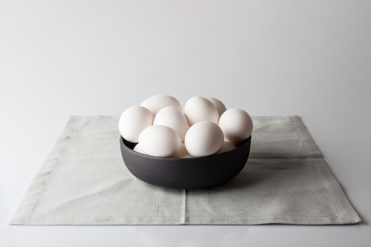 White eggs in a black bowl on beige napkin on white background from side