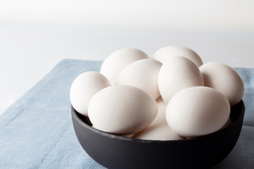 White eggs in a black bowl on blue linen napkin on white background from side off centre composition  