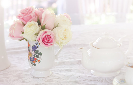 Beautiful soft pink and white roses in vase and teapot on table, covered with white cloth
