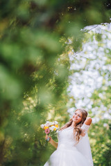 Gorgeous blonde bride in elegant dress holding bouquet and posing in the sunny summer park or garden on their wedding day