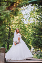 Obraz na płótnie Canvas Gorgeous blonde bride in elegant dress holding bouquet and posing in the sunny summer park or garden on their wedding day