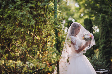 Gorgeous blonde bride in elegant dress holding bouquet and posing in the sunny summer park or garden on their wedding day