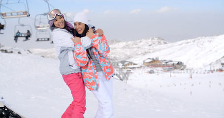 Two cute attractive young women at a ski resort standing below the ski lift hugging each other and laughing