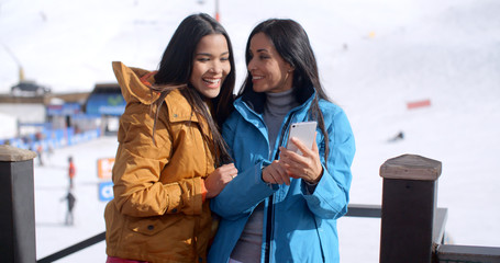 Two cute smiling young adult women in long black hair brown and blue jackets looking at a cell phone while standing in front of a ski slope.